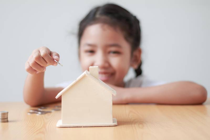 Asian little girl putting coin to house piggy bank shallow depth of field select focus on hand. Getty Images