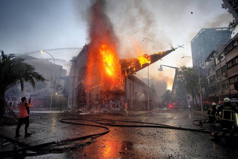 TOPSHOT - The dome of the church of Asuncion falls down burning in flames after being set on fire by demonstrators on the commemoration of the first anniversary of the social uprising in Chile, in Santiago, on October 18, 2020, as the country prepares for a landmark referendum. Two churches were torched as tens of thousands of demonstrators gathered Sunday in a central Santiago square to mark the anniversary of a protest movement that broke out last year demanding greater equality in Chile. The demonstration comes just a week before Chileans vote in a referendum on whether to replace the dictatorship-era constitution -- one of the key demands when the protest movement began on October 18, 2019. / AFP / CLAUDIO REYES
