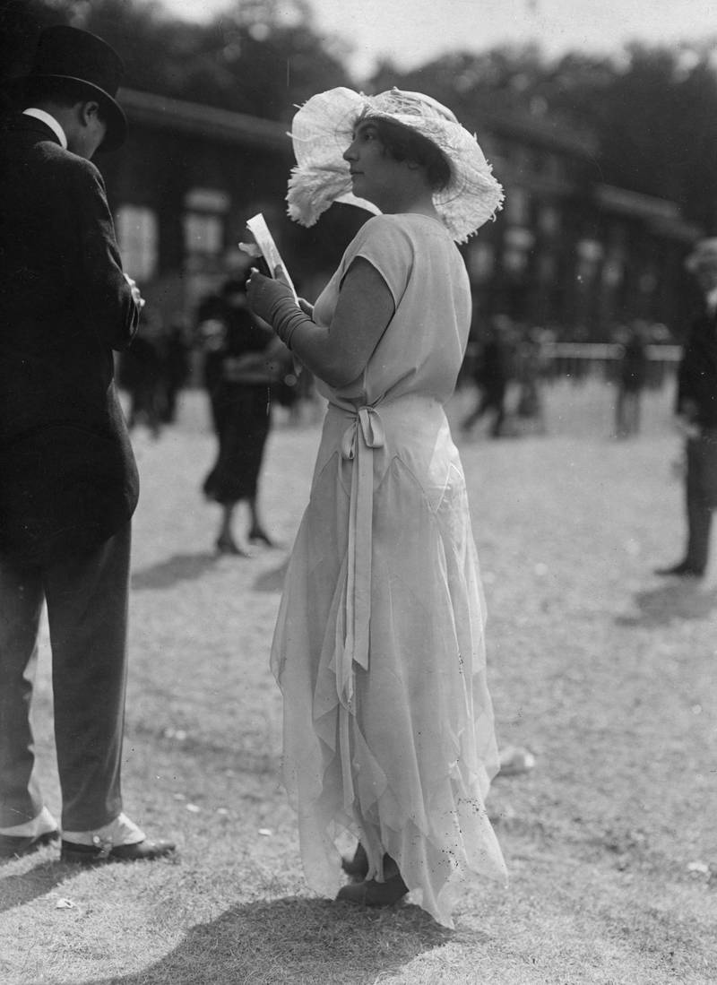 A women at Royal Ascot circa 1920 wears a white charmeuse and georgette Grecian dress, a wide-brimmed hat decorated with feathers and short gloves. Getty Images