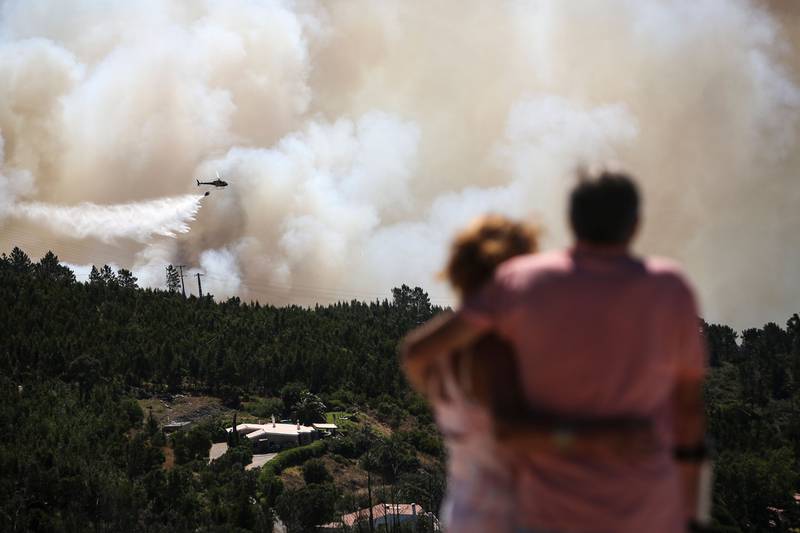 TOPSHOT - A couple watches an helicopter dropping water on a wildfire near Monchique, in Algarve, on August 8, 2018. - Spain and Portugal approached record temperatures at the weekend, with the mercury hitting 46.6 degrees Celsius (116 Fahrenheit) at El Granado in Spain and 46.4 C in Alvega, Portugal, according to the World Meteorological Organisation (WMO). While the deadly hot spell is expected to ease in parts of western Europe in the coming days, firefighters in Spain and Portugal struggled to contain wildfires that have swept southern areas. (Photo by CARLOS COSTA / AFP)