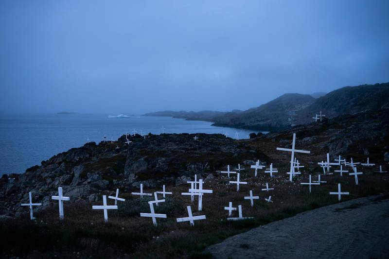 Crosses stand in a cemetery as an iceberg floats in the distance during a foggy morning.
