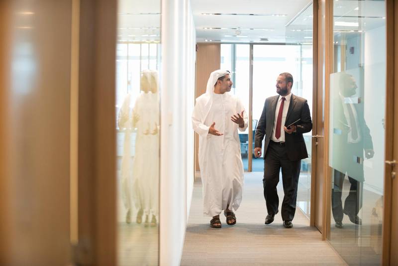 A photo of Middle Eastern Arab businessmen discussing in corridor. Full length of professionals are walking in modern office. One is in suit and other is wearing traditional Emirati clothes. Dubai, United Arab Emirates.