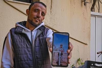 Fahad Al Routayban, 30, a Syrian refugee, poses for a picture the building where he works as a concierge, in Lebanon's northern port city of Tripoli on February 23, 2021, while holding a phone showing a photo of himself from 11 years prior as a Syrian army soldier.  Routayban fled his hometown of Raqa to Lebanon in 2013. He finally settled in Tripoli where he got married to a relative, another Syrian refugee, fathering two sons. AFP