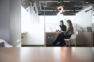 A photo of businesswomen holding document. Full length of multi-ethnic professionals are sitting at desk. They are discussing in cubicle, in brightly lit office. Caucasian and Middle Eastern Arab woman discuss document in Dubai office.