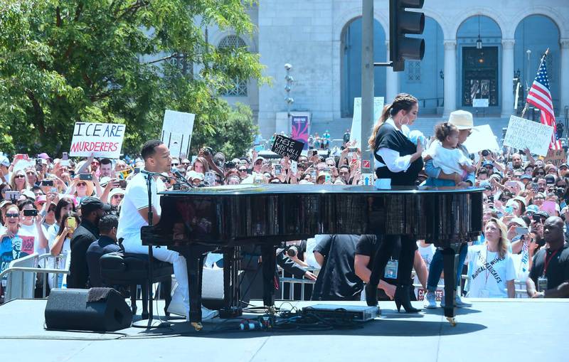 John Legend sings at the piano as Chrissy Teigen, holding their baby, walks off stage during a 'Familes Belong Together' march and rally in Los Angeles, California. AFP