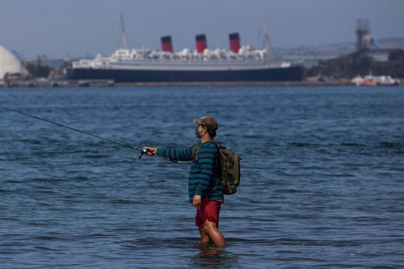 A man wearing a facemask fishes with the The Queen Mary ocean liner in the background in Long Beach, California, on July 14, 2020. California's Governor Gavin Newsom announced a significant rollback of the state's reopening plan on July 13, 2020 as coronavirus cases soared across America's richest and most populous state. / AFP / Apu GOMES
