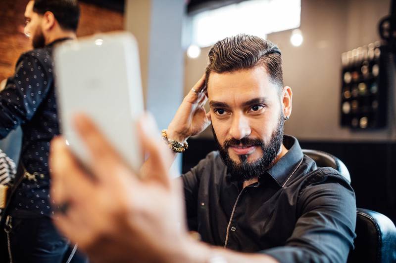 Male customer examining haircut on mobile phone. Satisfied client is touching hair in salon. Focus is on him. Getty Images