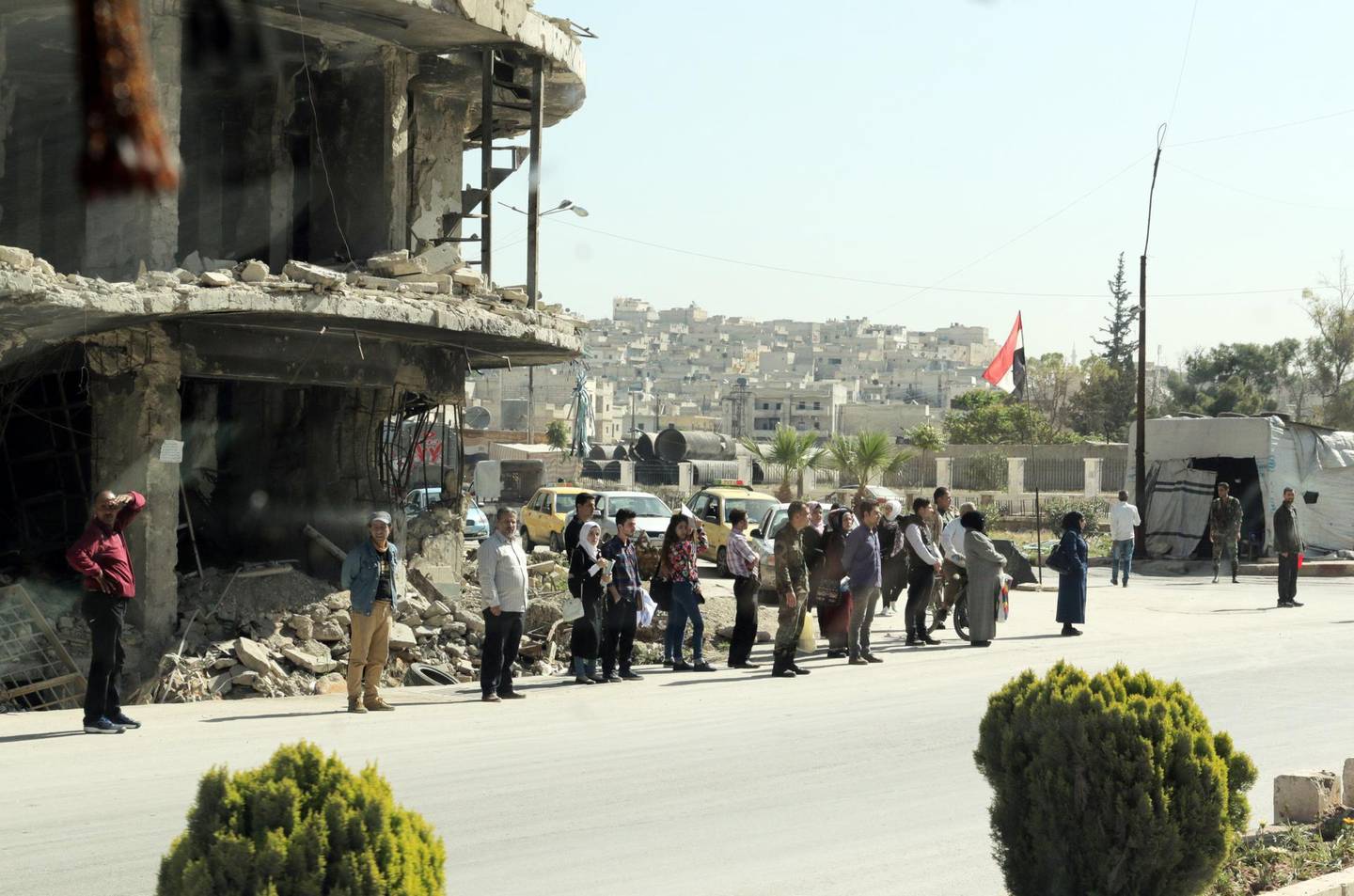 epa06677865 A picture taken through the window of a bus shows people standing in front of a destroyed building, in an unspecified town in Aleppo province, northern Syria, 18 April 2018. The previous day, folklore shows and traditional songs were performed in the Citadel Theater of besieged city of Aleppo to mark Syria's Independence Day.  EPA/YOUSSEF BADAWI