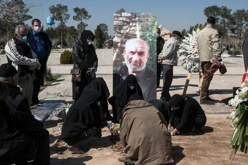 Relatives wearing face masks mourn over the grave of former politburo official in the Revolutionary Guard Farzad Tazari, shown in a poster, who died on Monday after being infected with the new coronavirus, at the Behesht-e-Zahra cemetery just outside Tehran, Iran.  AP