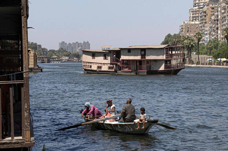 A family of fishermen sits in a boat as one of the houseboats (background) usually moored across one of the banks of the Nile river between the Zamalek district of Egypt's capital Cairo (R) and the Agouza district of its twin city of Giza (L) is towed away by authorities on June 27, 2022, as part of a wider decree to clear all of the river's banks in the area.  - Urban reprieve for some, life savings for others, around thirty Nile of the houseboats, known as "awamat" (floating), are slated for demolition in Cairo, with residents claiming the state is sacrificing heritage for profit.  A campaign to save the houseboats has been launched online, with a petition garnering more than 4,000 signatures.  The vessels hold cultural weight even beyond the Nile, cemented in Arab cinema as the sight where Abdel Halim Hafez crooned in 1955's 'Ayam w Layali' (Days and Nights) and the setting for the titular chitchat in 1971's 'Tharthara fawq al-Neel' (Chitchat on the Nile), based on the novel by Nobel Prize-winning Naguib Mahfouz.  (Photo by Khaled DESOUKI  /  AFP)