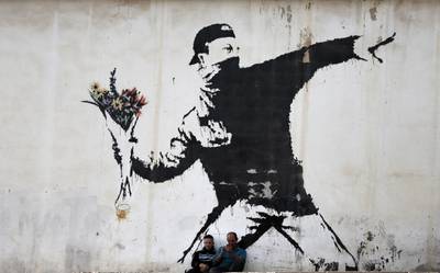 Two men are sitting in front of a famous graffiti of British street artist Banksy, painted on a wall of a gas station in the West Bank city of Bethlehem on December 16, 2015. AFP