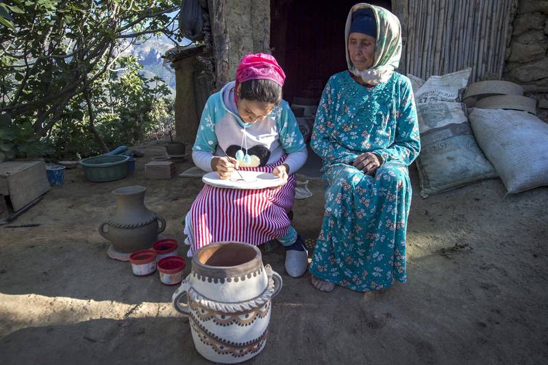 Moroccan potter Houda Oumal (L) paints with natural pigments on one of her pieces of pottery as her mother Fatima Harama looks on, near the village of Ourtzagh in the region of Taounate on june 11, 2019 The Sumano association, which promotes Moroccan tribal women's handicrafts, places orders with the potters, buys the works, transports them to Spain and sells them at 20 times the local price on its website, promising to redistribute the income locally "when the business becomes profitable". / AFP / FADEL SENNA
