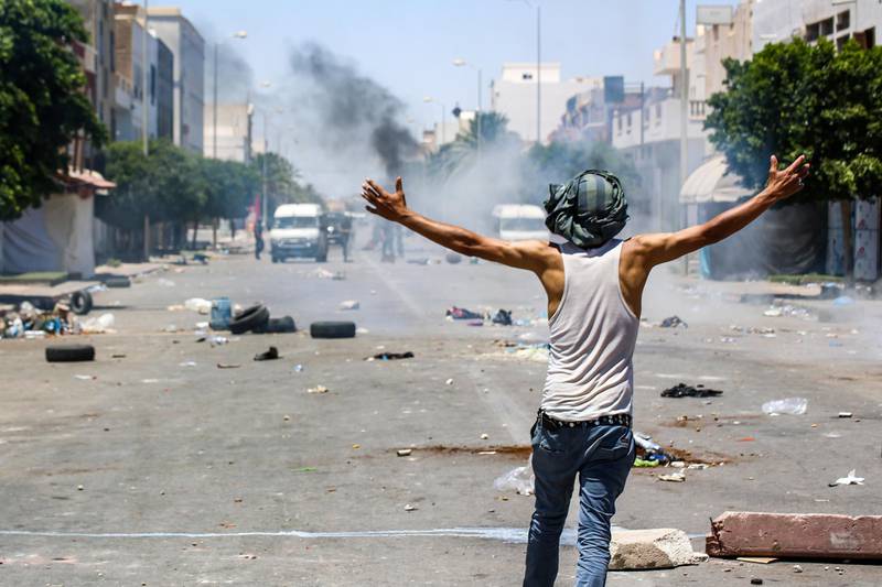A protester from Tunisia's Tataouine region walks with open arms as security forces stand across the street behind fumes during clashes amidst a demonstration in the southern city on June 21, 2020. Clashes erupted on June 21 in Tataouine in Tunisia between police and protesters demanding jobs in oil and gas companies, as well as the release of an activist. For several weeks, a protest movement has been underway in the southern province of Tataouine, where demonstrators have erected tents in several regions and blocked the road to trucks belonging to these companies located at the site of El Kamour, 160 kilometers, in the middle of the desert. / AFP / FATHI NASRI
