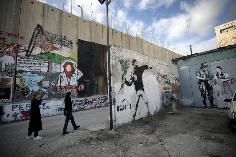 epa07226222 Tourists pass a graffiti of Banksy, anonymous England-based street artist (R) at the separation wall in the West Bank city of Bethlehem, 12 December 2108.  EPA-EFE/ATEF SAFADI *** Local Caption *** 54837282