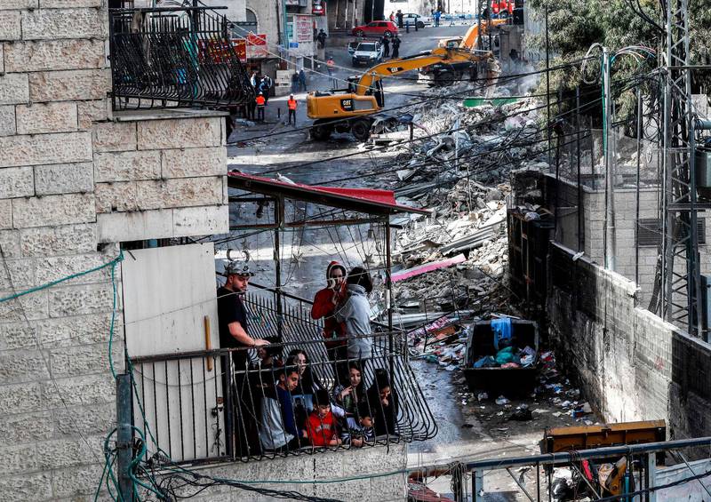 A Palestinian family watch from a balcony as excavators of the Jerusalem municipality demolish over a dozen shops in the Arab-inhabited Shuafat refugee camp in Israeli-annexed East Jerusalem on November 21, 2018, which authorities said were built without a permit.  / AFP / AHMAD GHARABLI
