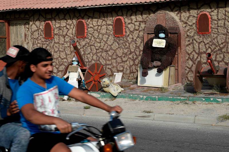 Youths ride a scooter past a home with a stuffed gorilla outside wearing a face mask with a sign reading in Arabic "I, the gorilla, wore a mask, why don't you wear (one)?", in Hamad Town, south of Bahrain's capital Manama on May 19, 2020.  AFP