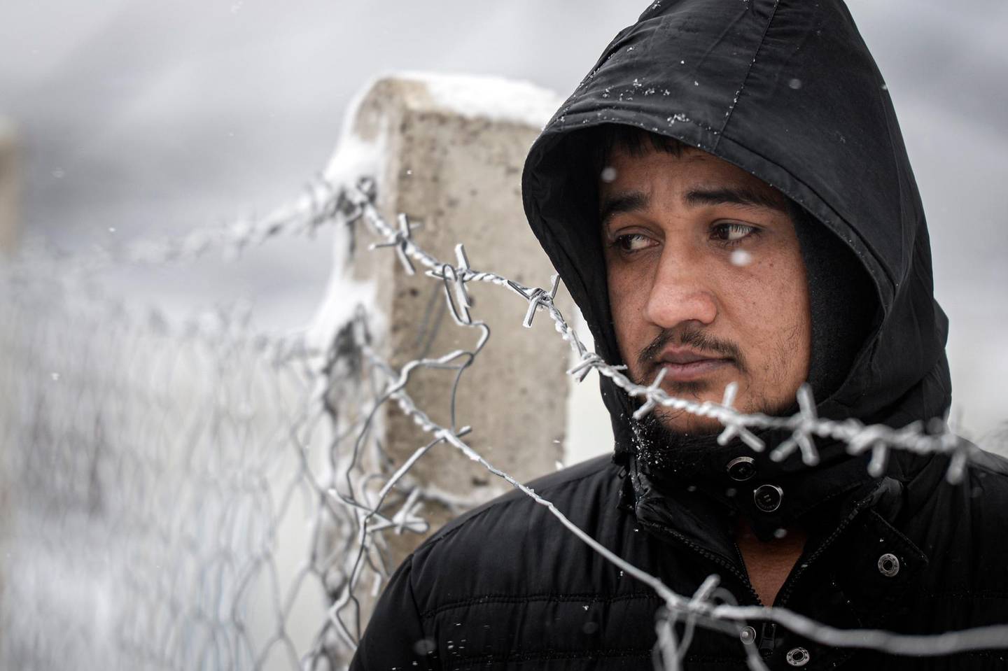 A migrant stands next to a fence during snowfall at the Lipa camp, outside Bihac, Bosnia, Friday, Jan. 8, 2021. A fresh spate of snowy and very cold winter weather on has brought more misery for hundreds of migrants who have been stuck for days in a burnt out camp in northwest Bosnia waiting for heating and other facilities. (AP Photo/Kemal Softic)