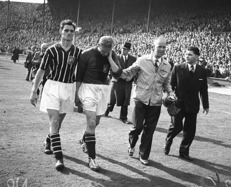 5th May 1956:  FA Cup Final: Birmingham City v Manchester City. After helping Manchester City win the Cup 3-1 Bert Trautmann, injured during the match, is helped off the field rubbing his 'sore'' neck. It later transpired he had broken it. Trautmann was later named as player of the year. Mandatory Credit: Allsport Hulton/Archive/Getty Images