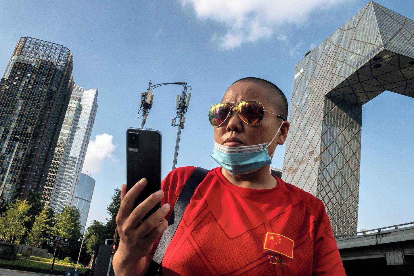 A woman checks her smartphone as she walks before two cellphone towers (back C), used for a 5G network, on a street in Beijing on September 24, 2020. (Photo by NICOLAS ASFOURI / AFP)