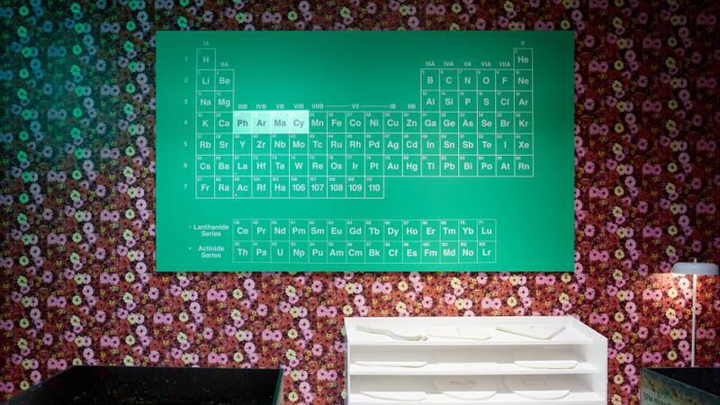 A Hirst artwork based on the periodic table, which has been altered to read pharmacy. It is placed over wallpaper decorated with flowers and cigarette butts.
