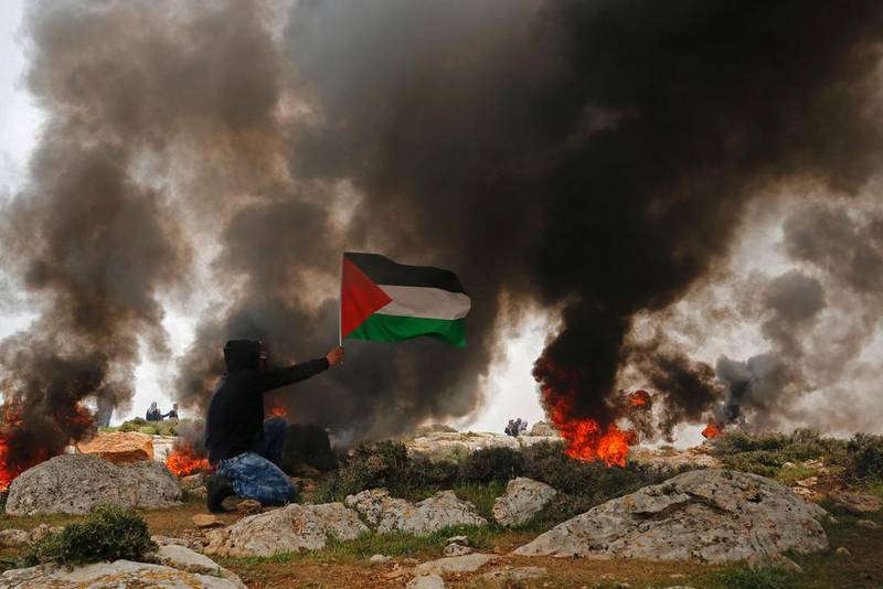 A Palestinian protester holds his national flag amidst smoke from burning tyres following a demonstration against Jewish settlements in the village of Al Mughayyir near the West Bank city of Ramallah on March 24, 2017. Abbas Momani / Agence France-Presse