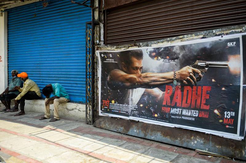 People sit in front of a closed shop next to a poster of the upcoming Bollywood movie 'Radhe,' scheduled to be released on May 13 amidst the Covid-19 coronavirus pandemic, in Secunderabad, the twin city of Hyderabad, on May 11, 2021. (Photo by Noah SEELAM / AFP)