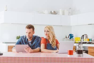 Adult couple after argument, sitting at the table in the kitchen at home. Man using a digital tabet and woman typing on laptop, peeking at her partner.