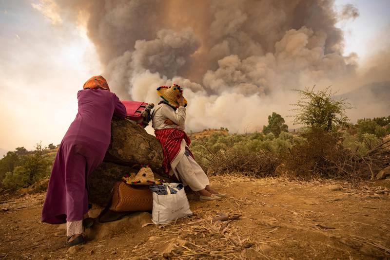 A woman stands on a mountainous side road with her belongings as wildfires spread across Morocco.