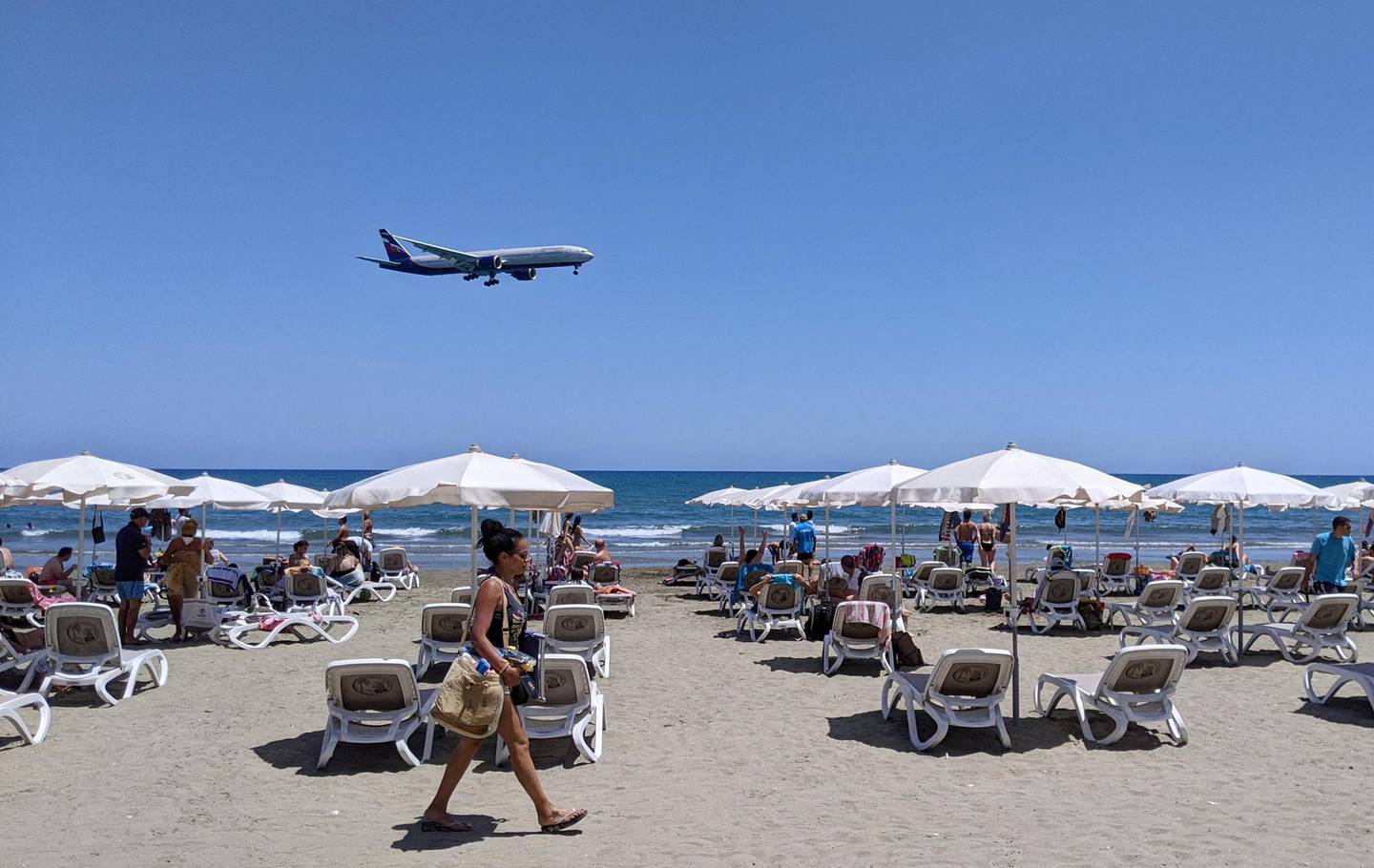 A passenger plane approaches Larnaca airport before landing as a woman walks past sunbeds at Makenzy Beach in the southern Cypriot port city on May 23, 2021. A 39-year-old British woman died in a Cypriot hospital as a result of  a blood clotting incident after receiving the AstraZeneca Covid-19 vaccine, the official Cyprus News Agency said May 24, 2021. / AFP / Etienne TORBEY
