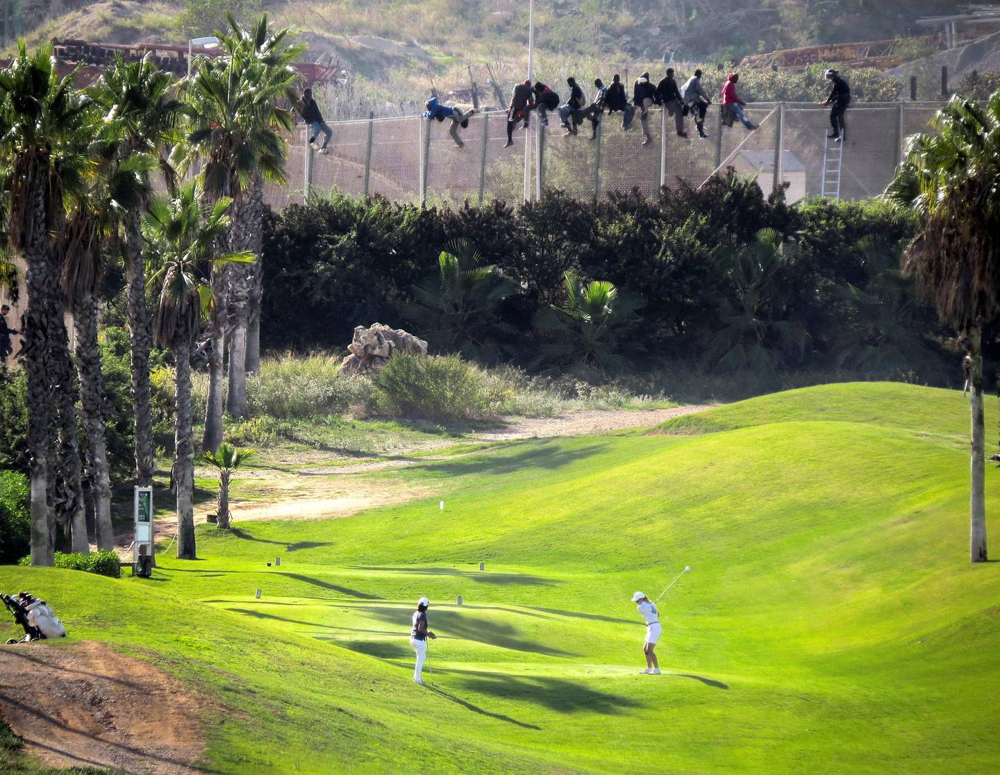 A golfer hits a tee shot as African migrants sit atop a border fence during an attempt to cross into Spanish territories between Morocco and Spain's north African enclave of Melilla October 22, 2014. Around 400 migrants attempted to cross the border into Spain, according to local media. Picture taken October 22, 2014. REUTERS/Jose Palazon  (SPAIN - Tags: SPORT GOLF SOCIETY IMMIGRATION TPX IMAGES OF THE DAY)  BEST QUALITY AVAILABLE - GM1EAAN1CDU01