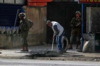 A Palestinian man uses a stick to remove burnt tyres left in in front of his house as Israeli soldiers stand guard. EPA