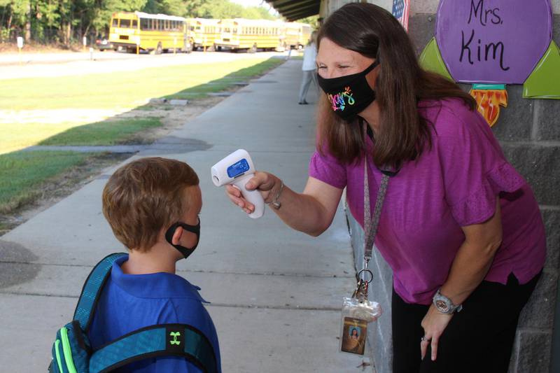 In a photo provided by Newton County Schools, teaching assistant Crystal May talks to kindergarten student Lewis Henry Thompson, 5, as she takes his temperature at Newton County Elementary School in Decatur, Miss., Monday, Aug. 3, 2020. Thousands of students across the nation resumed in-person school Monday for the first time since March. Parents are having to balance the children's need for socialization and instruction that school provides, with the reality that the U.S. death toll from the coronavirus has hit about 155,000 and cases are rising in numerous places. (Janine Vincent/Newton County Schools via AP)