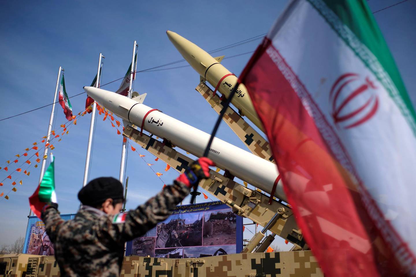 An Iranian young boy wearing an Islamic Revolutionary Guard Corps (IRGC) uniform holds an Iran flag while Iran-made, Dezful medium range ballistic missile (Bottom) and Zolfaghar road-mobile single-stage solid-propelled liquid fueled missile are pictured in the Azadi (Freedom) square during a rally to commemorate the 42nd Victory anniversary of the Islamic Revolution, that held with motorcycles amid the new coronavirus disease (COVID-19) outbreak in Iran, in Tehran on February 10, 2021, on February 10, 2021.  (Photo by Morteza Nikoubazl/NurPhoto via Getty Images)