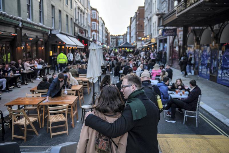 LONDON, ENGLAND - APRIL 12: A couple embrace in front of a street of people eating and drinking in Soho as non essential retail reopens on April 12, 2021 in London, United Kingdom. England has taken a significant step in easing its lockdown restrictions, with non-essential retail, beauty services, gyms and outdoor entertainment venues among the businesses given the green light to re-open with coronavirus precautions in place. Pubs and restaurants are also allowed open their outdoor areas, with no requirements for patrons to order food when buying alcoholic drinks.  (Photo by Chris J Ratcliffe/Getty Images)