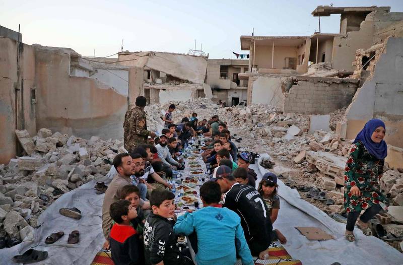 This picture taken on May 12, 2020 in the village of Kafr Nuran in the western countryside of Syria's northern Aleppo province shows men and children seated together in the midst of ruins before starting the "iftar" fast-breaking meal consumed by Muslims at sunset during the holy fasting month of Ramadan, as part of efforts by the humanitarian non-governmental organisation Caravanes Solidaires.  / AFP / Aaref WATAD
