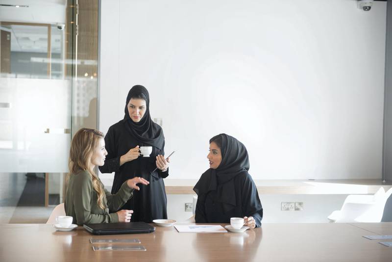 A photo of multi-ethnic businesswomen discussing. Arab Emirati women are in traditional abaya clothes and Caucasian female is in western dress. Professionals are at conference table, in brightly lit modern office discussing business cooperation. Dubai, United Arab Emirates.