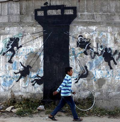 epa04699335 A Palestinian boy walks past a graffiti mural of children using an Israeli army watch tower as a swing ride, presumably painted by British street artist Banksy, on the wall of destroyed homes in Beit hanun town in the northern Gaza Strip, 10 April 2015.  EPA/MOHAMMED SABER *** Local Caption *** 51882350