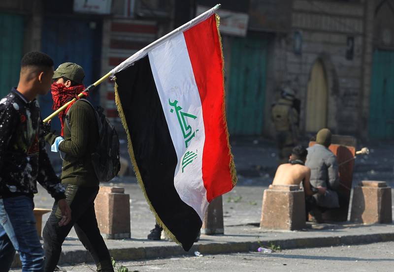 An Iraqi protester carries a national flag as others take cover behind a makeshift shield amid clashes with riot police following a demonstration at Al Wathba Square in the capital Baghdad, on January 30, 2020. Iraq's political factions were in high-stake talks to name a new prime minister ahead of a February 1 deadline to replace current caretaker Adel Abdel Mahdi who resigned in December, after two months of deadly protests in Iraq's capital and Shiite-majority south expressing outrage at rampant corruption and unemployment, quickly escalating to calls for the government to resign. / AFP / AHMAD AL-RUBAYE
