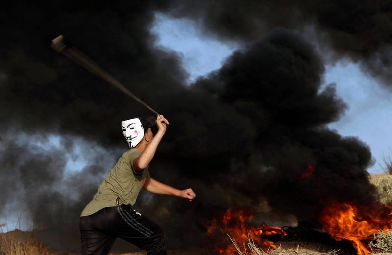 A protester wearing a mask hurls stones while others burn tyres near the fence of the border. AP Photo