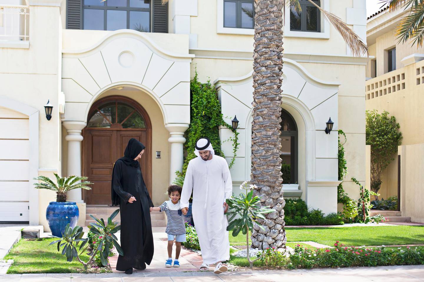 Happy middle Eastern family walking in the front garden of their house on a sunny day, both parents wearing the traditional Emirati dresses .