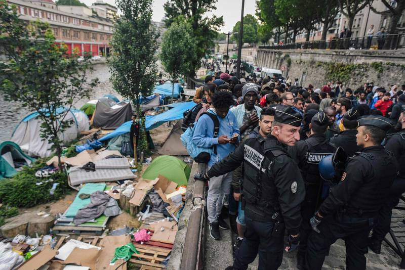 TOPSHOT - French CRS anti-riot police officers stand guard by migrants and refugees queing during the evacuation by police of their makeshift camp along the Canal de Saint-Martin at Quai de Valmy in Paris, on June 4, 2018. More than 500 migrants and refugees were evacuated on early June 4, 2018 from a makeshift camp that had been set up for several weeks along the Canal. / AFP / LUCAS BARIOULET
