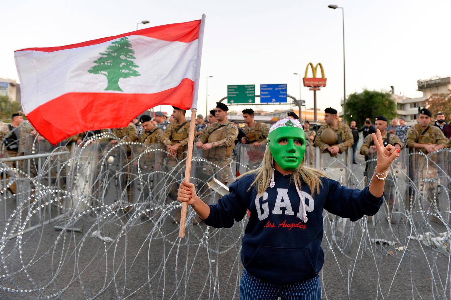 epa08027258 A protester wears a mask and holds a national flag as other protesters block the highway leading to the Presidential palace during a protest to demand the formation of a new government in Baabda, east Beirut, Lebanon, 26 November 2019. Protests in Lebanon are continuing since first erupted on 17 October, as protesters aim to apply pressure on the country's political leaders over what they view as a lack of progress following the prime minister's resignation on 29 October.  EPA/WAEL HAMZEH