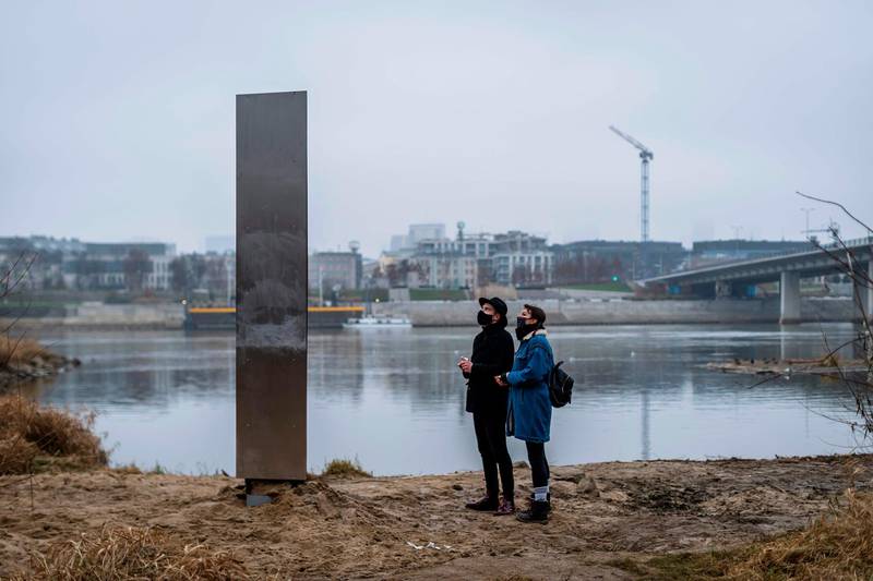 People look on December 10, 2020 at a metal monolith that has popped up on a riverbank of the Vistula in the Polish capital Warsaw, the latest in a string of similar objects that have recently appeared in Europe and the US. Similar-looking objects were found in the United States, Romania and Britain. / AFP / Wojtek RADWANSKI
