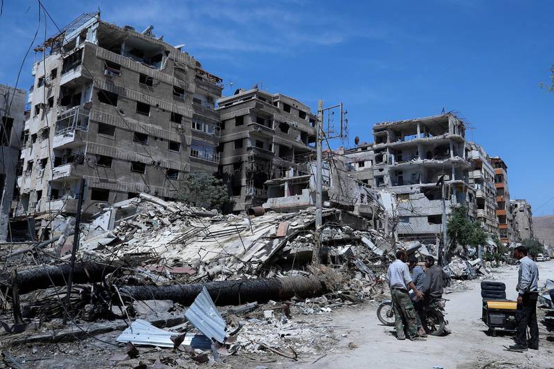 FILE - In this April 16, 2018, file photo, people stand in front of damaged buildings, in the town of Douma, near Damascus, Syria. In its ninth year, many Syrians fear their countryâ€™s unresolved war has become a footnote in a long list of world crises. (AP Photo/Hassan Ammar, File)