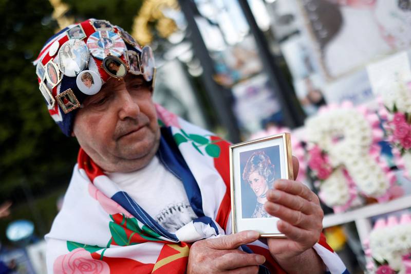 A man wrapped in the British national flag and wearing a hat covered with pins of the British royal family holds a photo portrait of Britain's Princess Diana during a gathering in front of Kensington Palace, in central London, on August 31, 2022 on the 25th anniversary of the Princess of Wales's death.  - Flowers and other tributes were laid on August 31, 2022 at princess Diana's former London home and above the Paris road tunnel where she lost her life, to mark the 25th anniversary of her death.  The former Lady Diana Spencer, whose fairytale marriage to Prince Charles captivated the world until it publicly unravelled with infidelity and divorce, died in a car crash in the French capital on August 31, 1997.  (Photo by CARLOS JASSO  /  AFP)