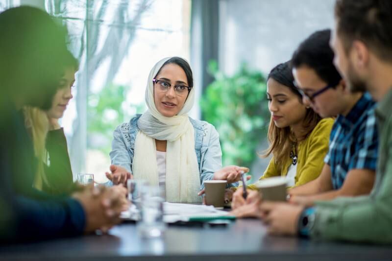A multi-ethnic group of business people are indoors in an office boardroom. They are sitting around a table and making plans. A woman wearing a head scarf is explaining papers to her coworkers.