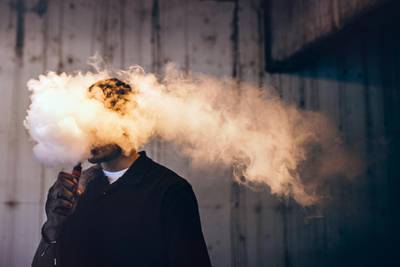 A dark and high contrast image of a young adult male has a cloud of nicotine vapor "smoke" pouring from his mouth.  Vape smoking, or "vaping" is growing in popularity, as well as falling under stricter state and governmental regulations.  Horizontal image with copy space.
