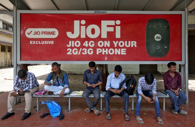 FILE PHOTO: Commuters use their mobile phones as they wait at a bus stop with an advertisement of Reliance Industries' Jio telecoms unit, in Mumbai, India July 10, 2017. REUTERS/Shailesh Andrade/File Photo