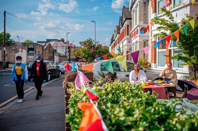 People wearing protective face masks walk past Walthamstow residents having a street party in their front garden to celebrate the 75th anniversary of VE Day (Victory in Europe Day), the end of the Second World War in Europe in Walthamstow in East London on May 8, 2020. / AFP / Tolga AKMEN
