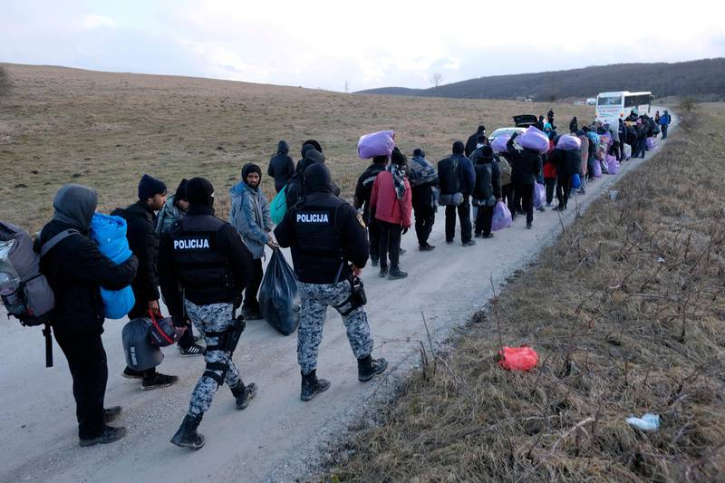 Police officers walk by migrants as they leave to be relocated from the Lipa camp. AP Photo